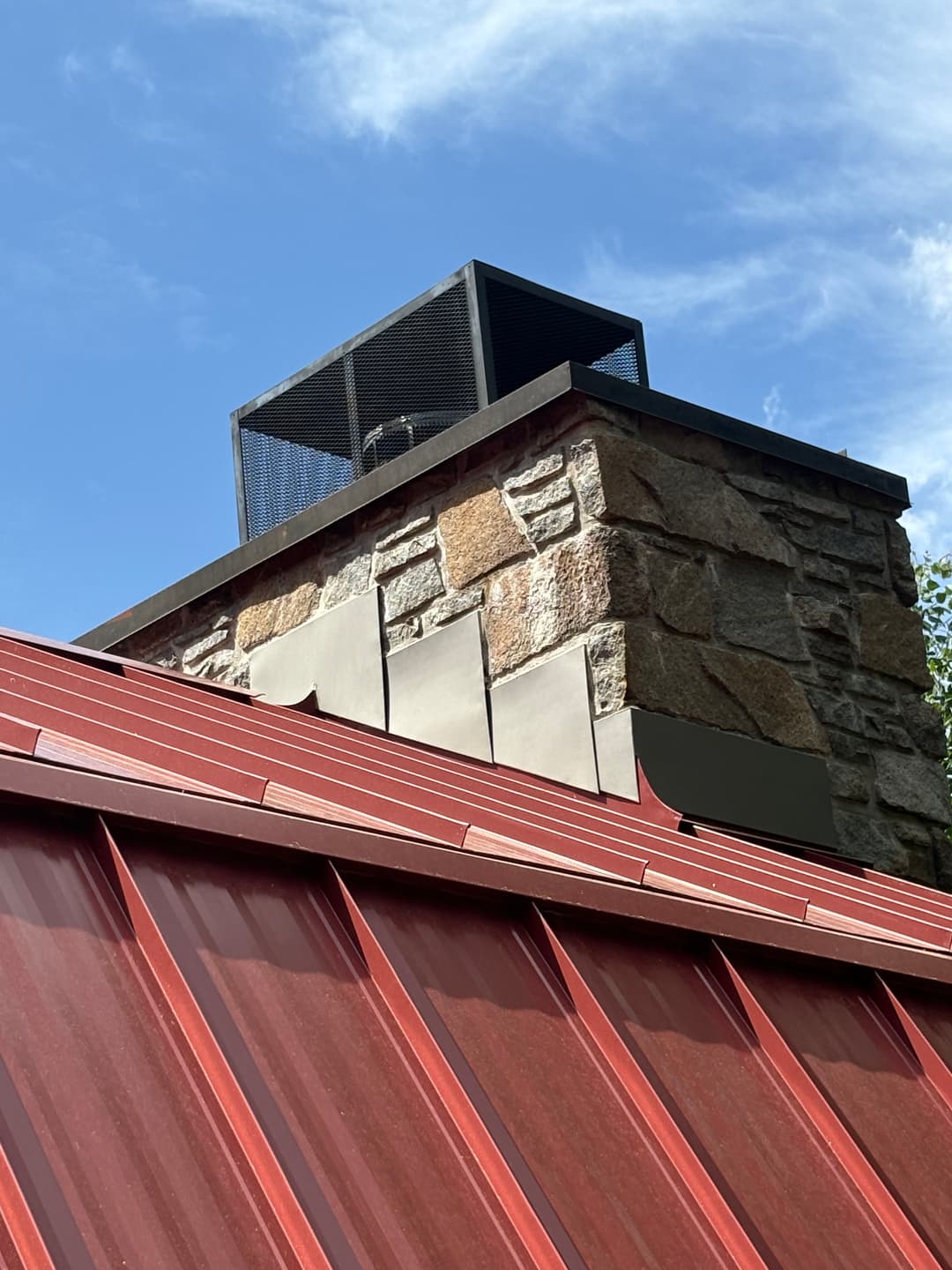 Chimney atop a stone structure with a metal cap against a blue sky. Red metal roof below.