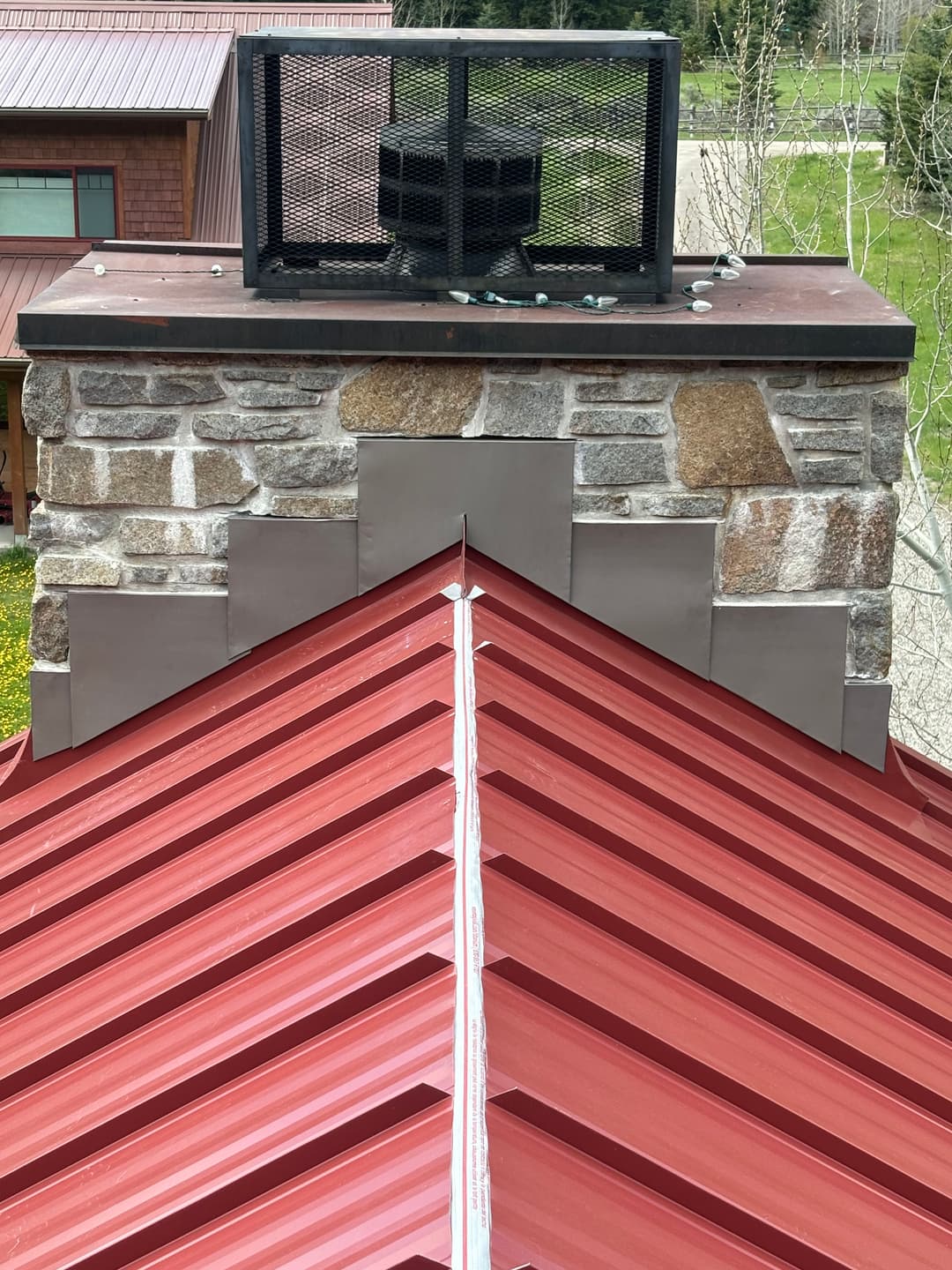 Red metal roof with stone chimney and ventilation fan in a residential setting.