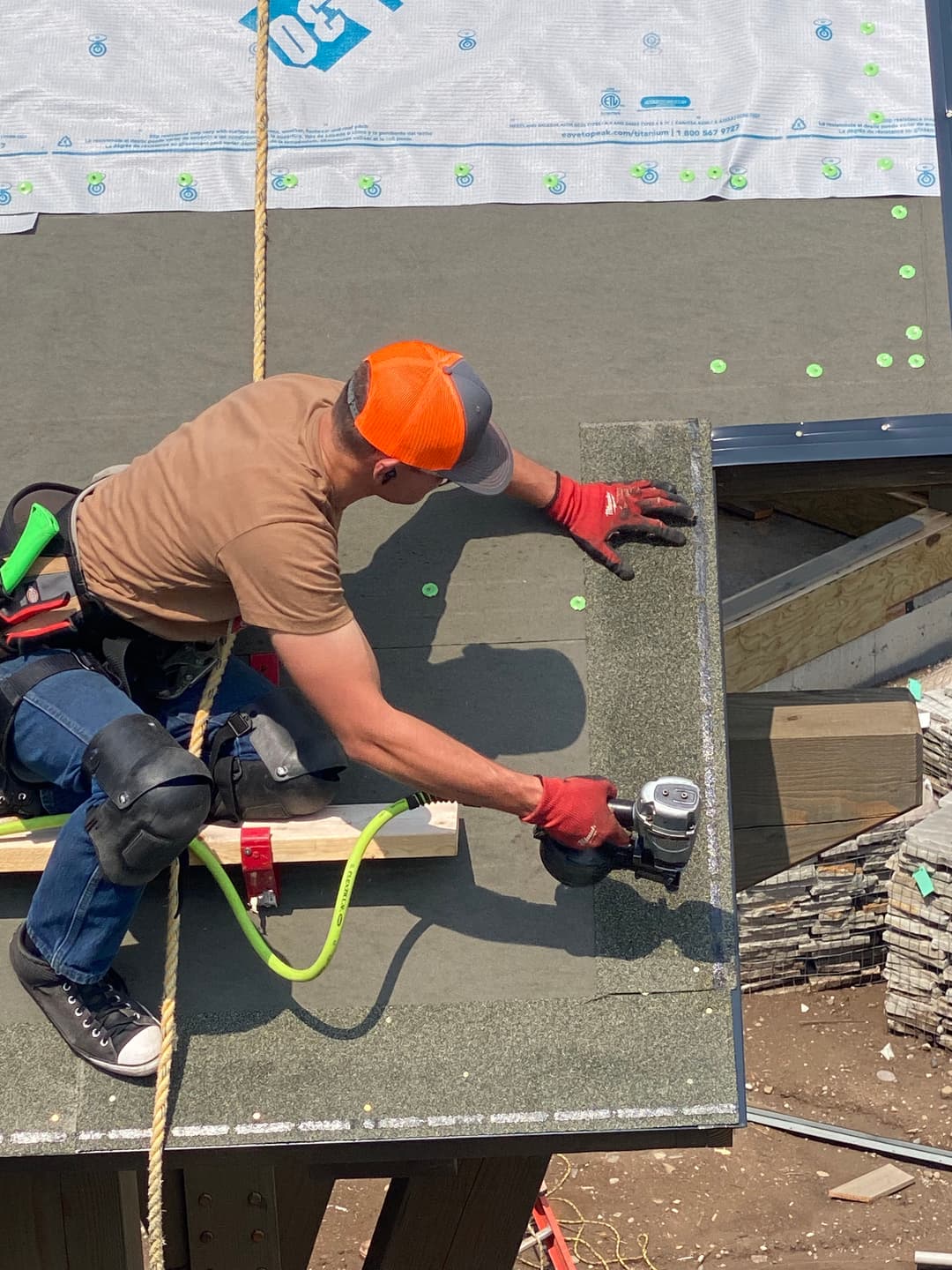 Roofer installing shingles on a sloped roof with tools and safety gear.
