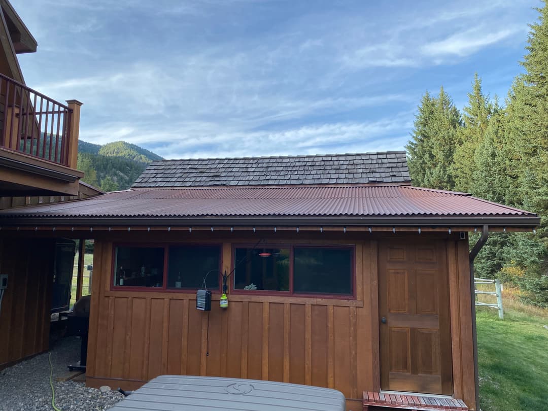 Wooden cabin with red roof, surrounded by trees and mountains under a clear blue sky.