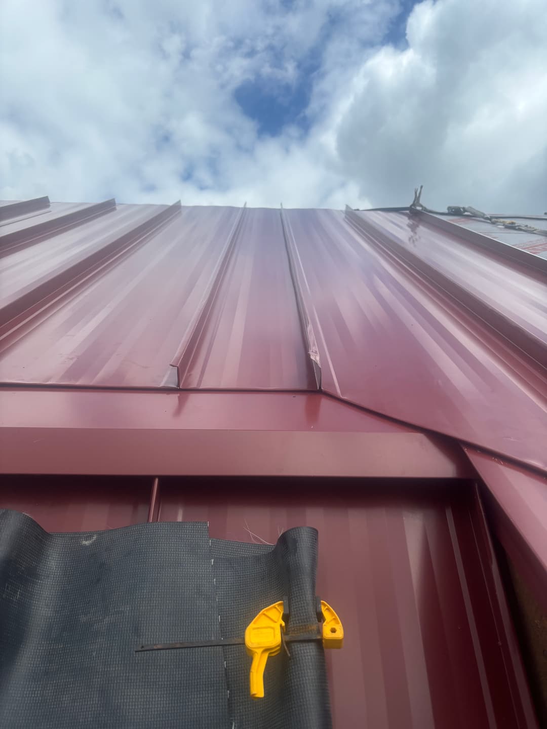 Close-up of a red metal roof with a yellow measuring tool and partly cloudy sky above.
