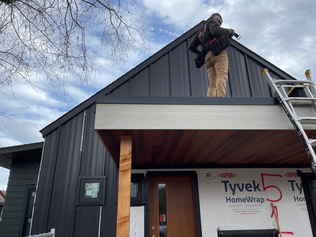 Worker installing roofing on a black metal home with Tyvek wrapping and wooden porch.