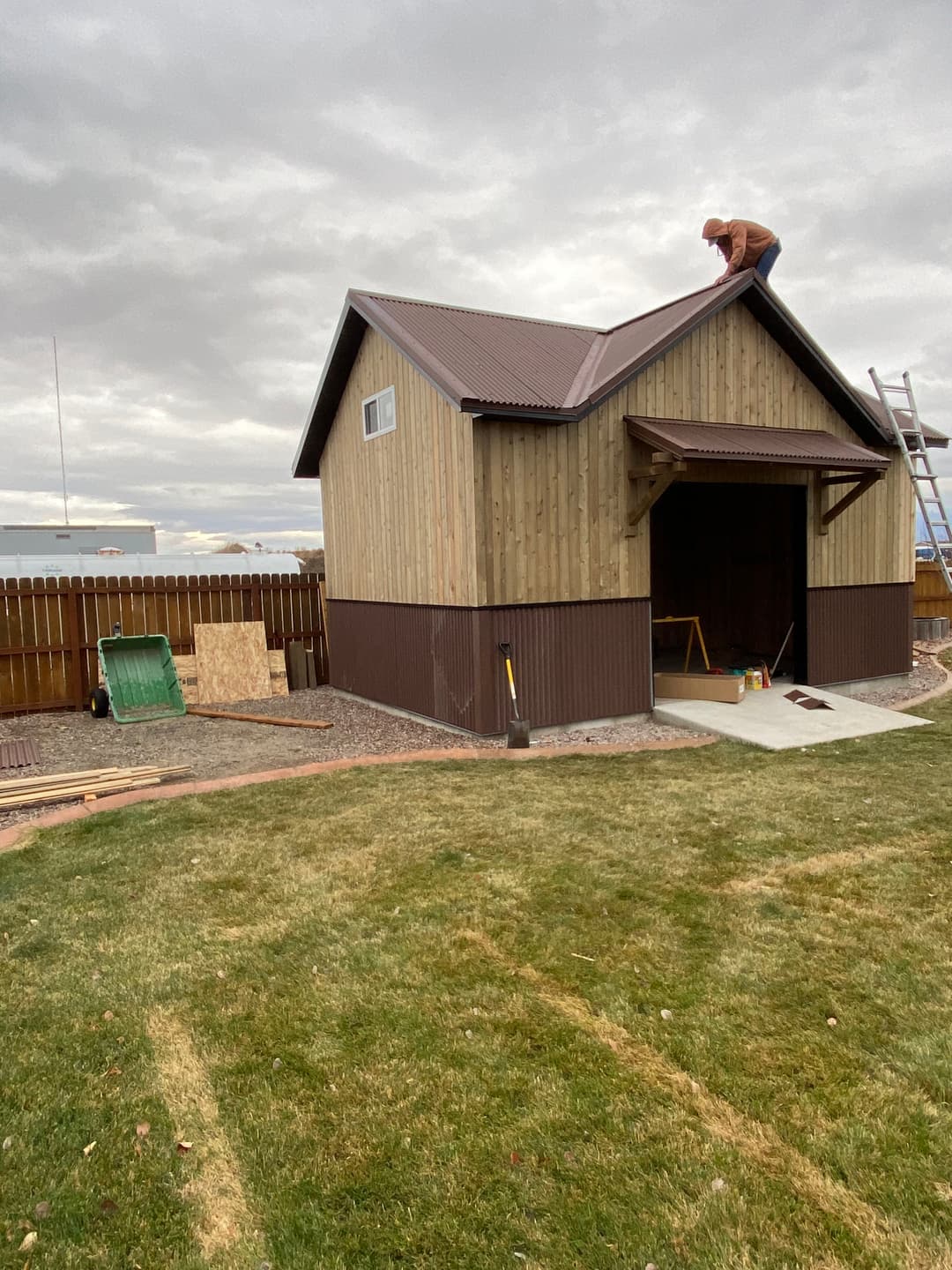 Person installing roofing on a wooden shed with a grassy yard and tools nearby.