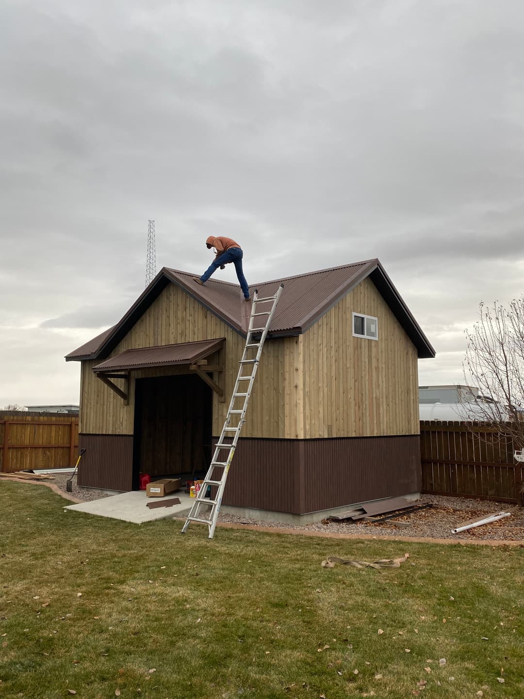 Man on ladder installing roofing on a wooden shed under cloudy sky.