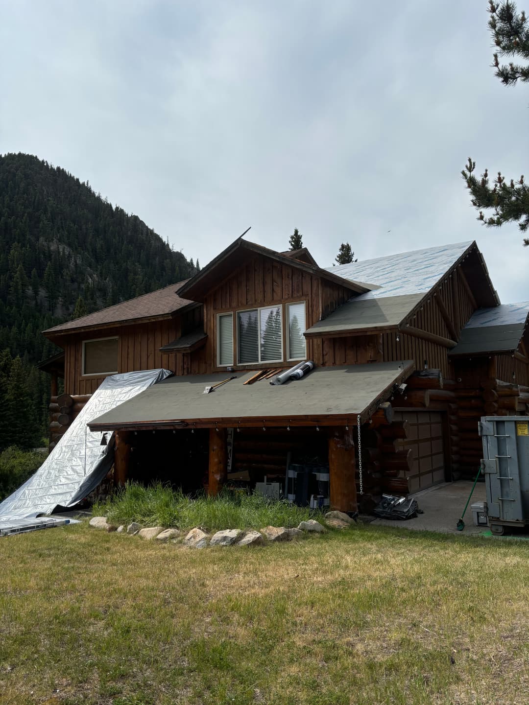 Log cabin home under renovation with tarps, mountains in the background, and green grass.