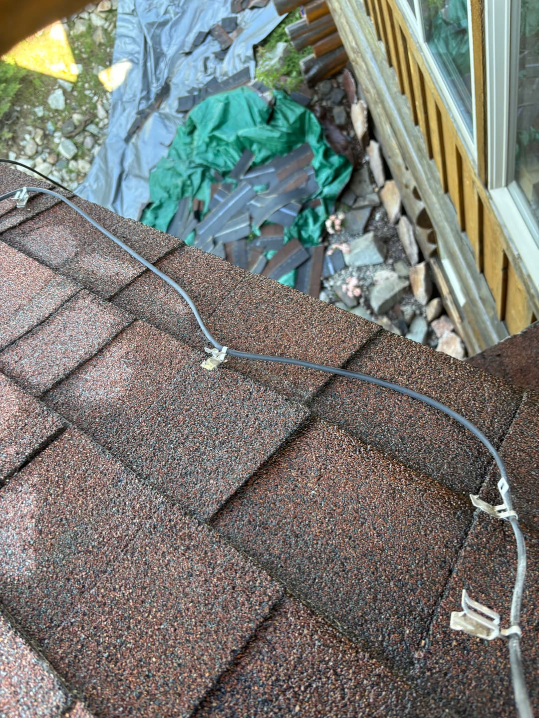 Electrical wiring on a roof shingles with green tarps and stone in the background.
