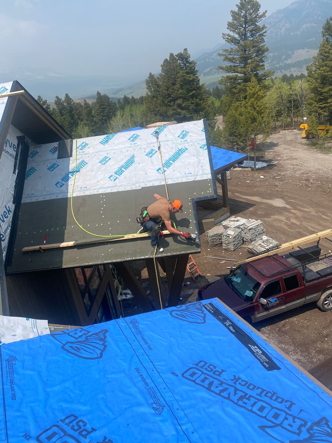 Construction worker installing roofing on a mountainside home under a clear blue sky.