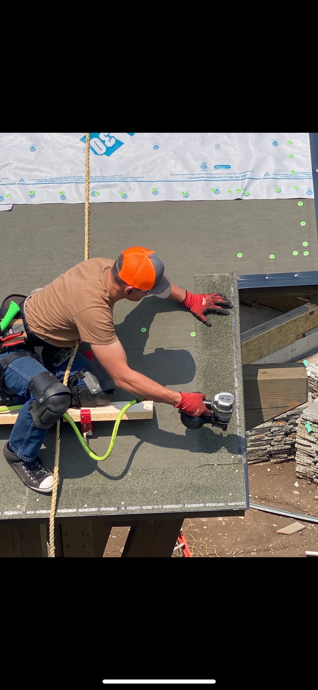 Contractor installing roofing materials with a nail gun on construction site. Safety gear visible.