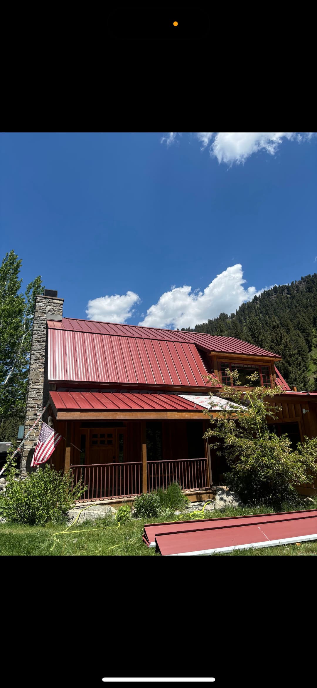 Red roofed mountain house with stone accents, surrounded by lush greenery and blue sky.
