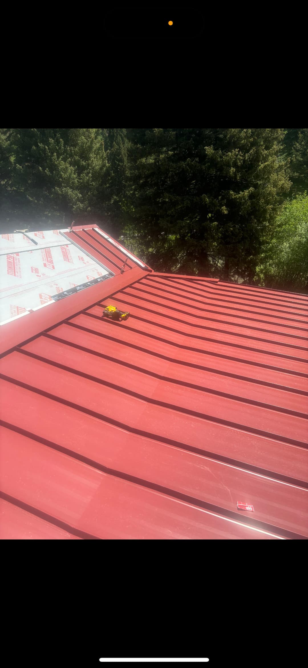 Red metal roof installation with bright yellow tool on sloped surface under clear blue sky.