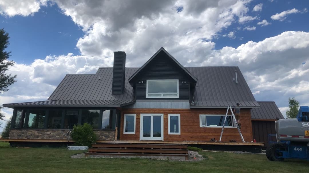 Modern home with a metal roof and large windows, surrounded by green grass and dramatic clouds.