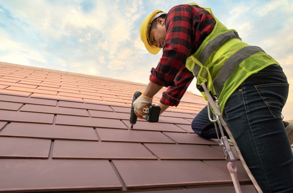 Construction worker installing roof tiles with a drill, wearing a safety vest and helmet.